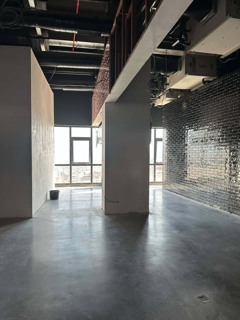 Empty industrial loft space with concrete floors, white drywall columns, exposed brick wall, and large windows with city view